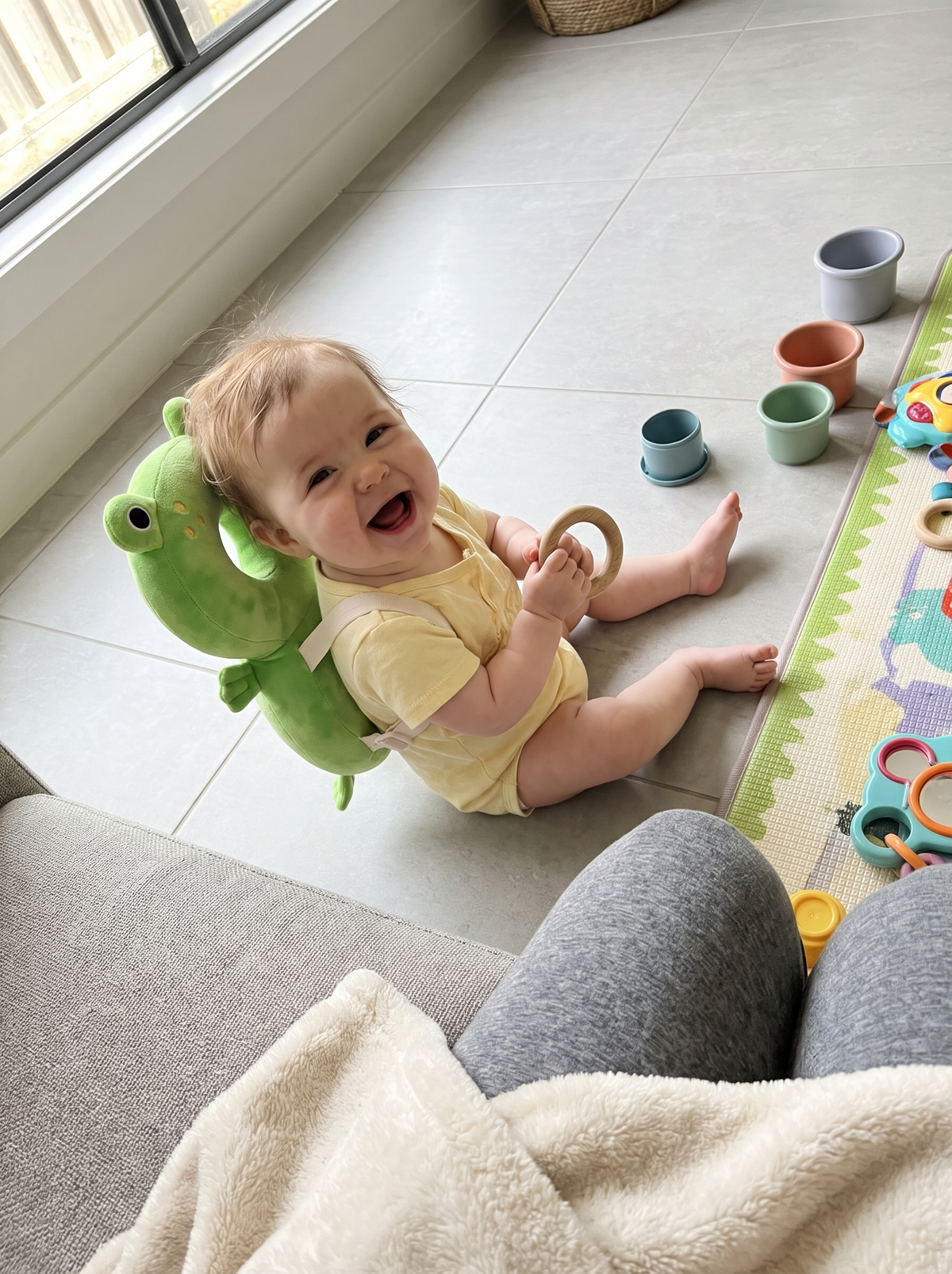 Baby girl wearing the frog head cushion backpack, sitting on a tiled floor