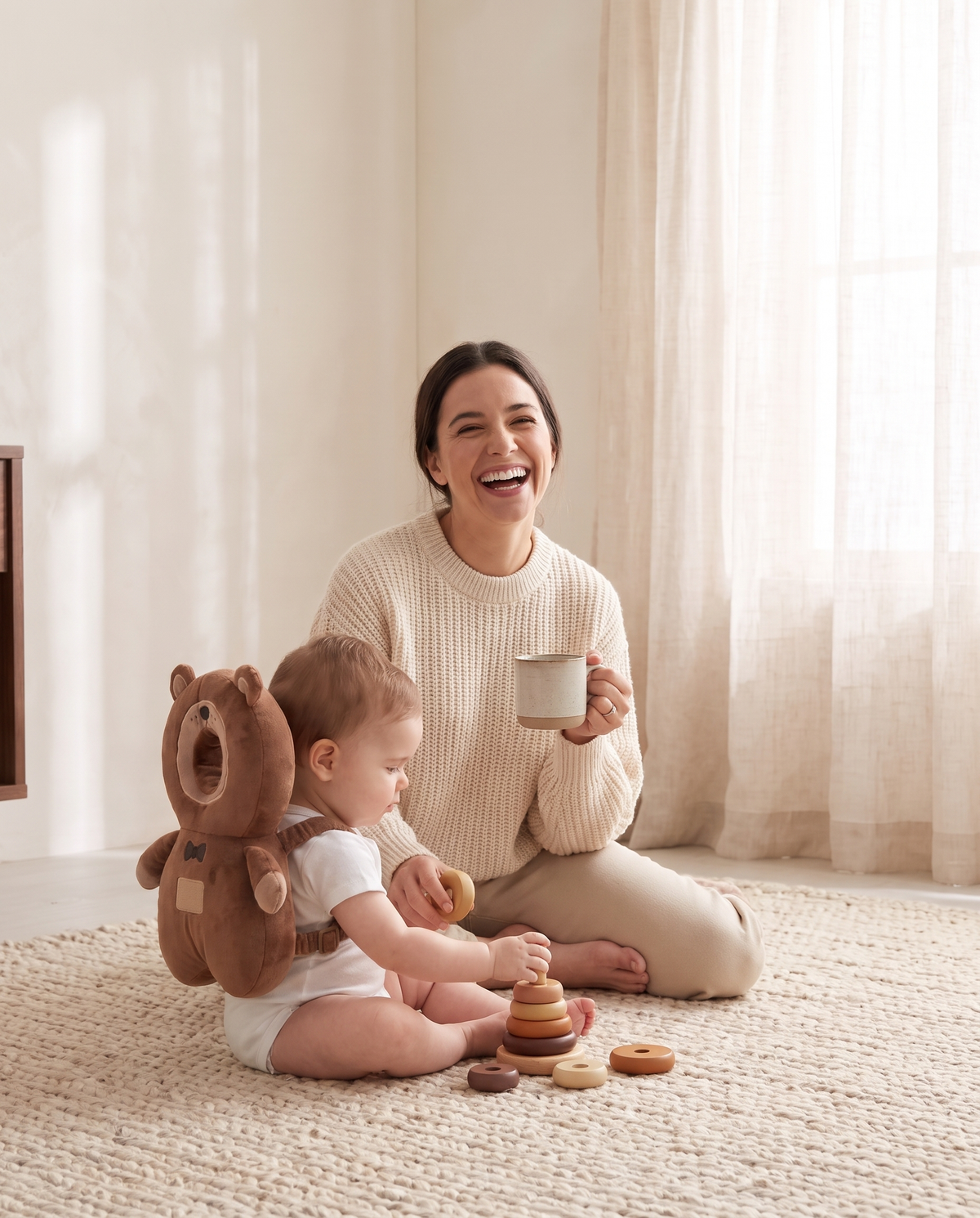 Mother laughing on the rug while her baby plays wearing a plush bear head protector backpack