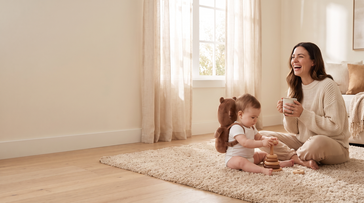 Mother laughing on the living room rug while her baby plays wearing a plush bear head protector backpack